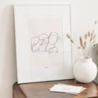 Tableau personnalisé couple dessin minimaliste sur fond uni posé sur un meuble en bois avec vase décoratif. Un portrait de couple dessiné au trait par Denadda dans le style line art.